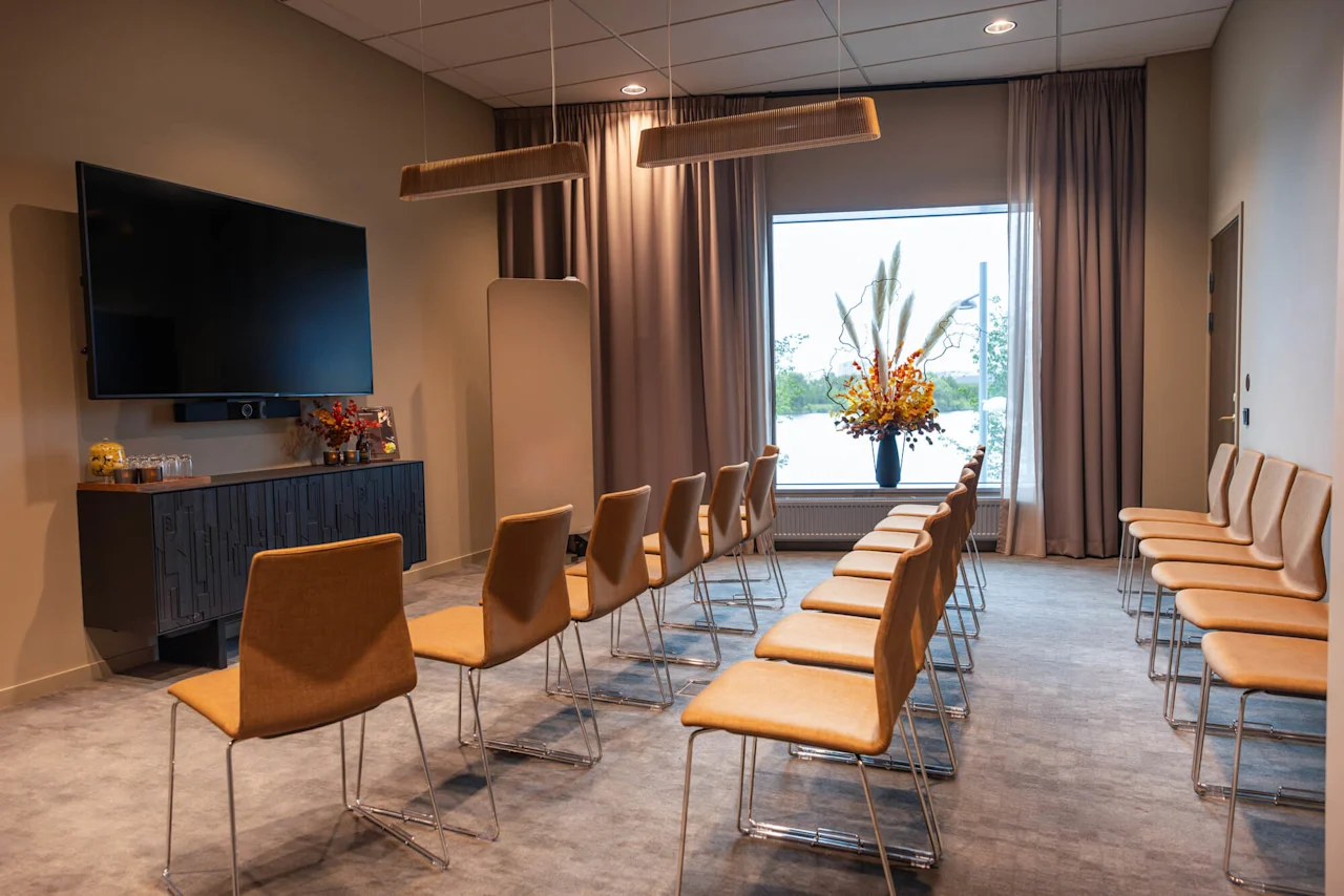 Rækker af brune stole står mod en stor vægmonteret TV i et moderne konferencelokale, forsynet med elegante loftslamper og et vindue med udsigt over en naturskøn udsigt og et blomsterarrangement.