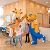 Two costumed mascots, a giraffe and a bear, high-five children in a wood-paneled room. The bear's outfit reads "Lollo & Bernie." Bright interior lighting enhances the lively atmosphere.
