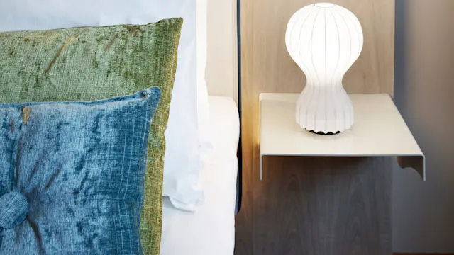 A decorative sheep head with glasses mounted on a wall above a bed, featuring green and blue pillows and a white lamp on a wooden nightstand.