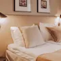 A neatly made bed with beige pillows and a matching bedspread; wall-mounted lamps illuminate framed pictures above, in a softly lit modern bedroom.