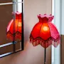 En röd lampskärm med dekorativ frans reflekteras i en väggmonterad spegel, bredvid ett tänt ljus på en vit hållare, vilket skapar en varm stämning.