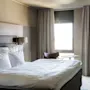 Bed with white linens placed against the left wall; light spills through a window, surrounded by beige curtains. A chair, small table, and floor lamp occupy the room.