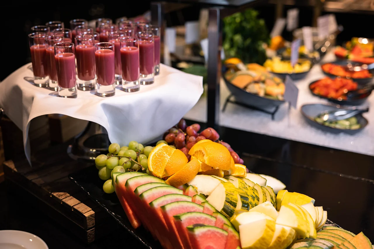 Glass fulle av lilla smoothie står på et dukdekket brett, med en fargerik fruktfat foran og flere retter i buffébaksiden.