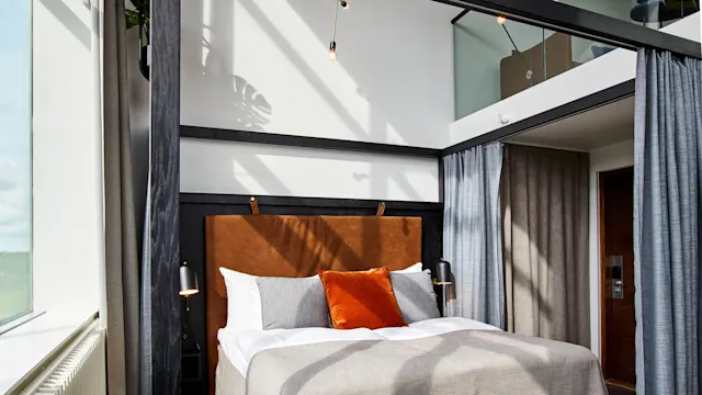 En säng med orange och grå kuddar står under hänglampor i ett rum med höga fönster och grå gardiner.