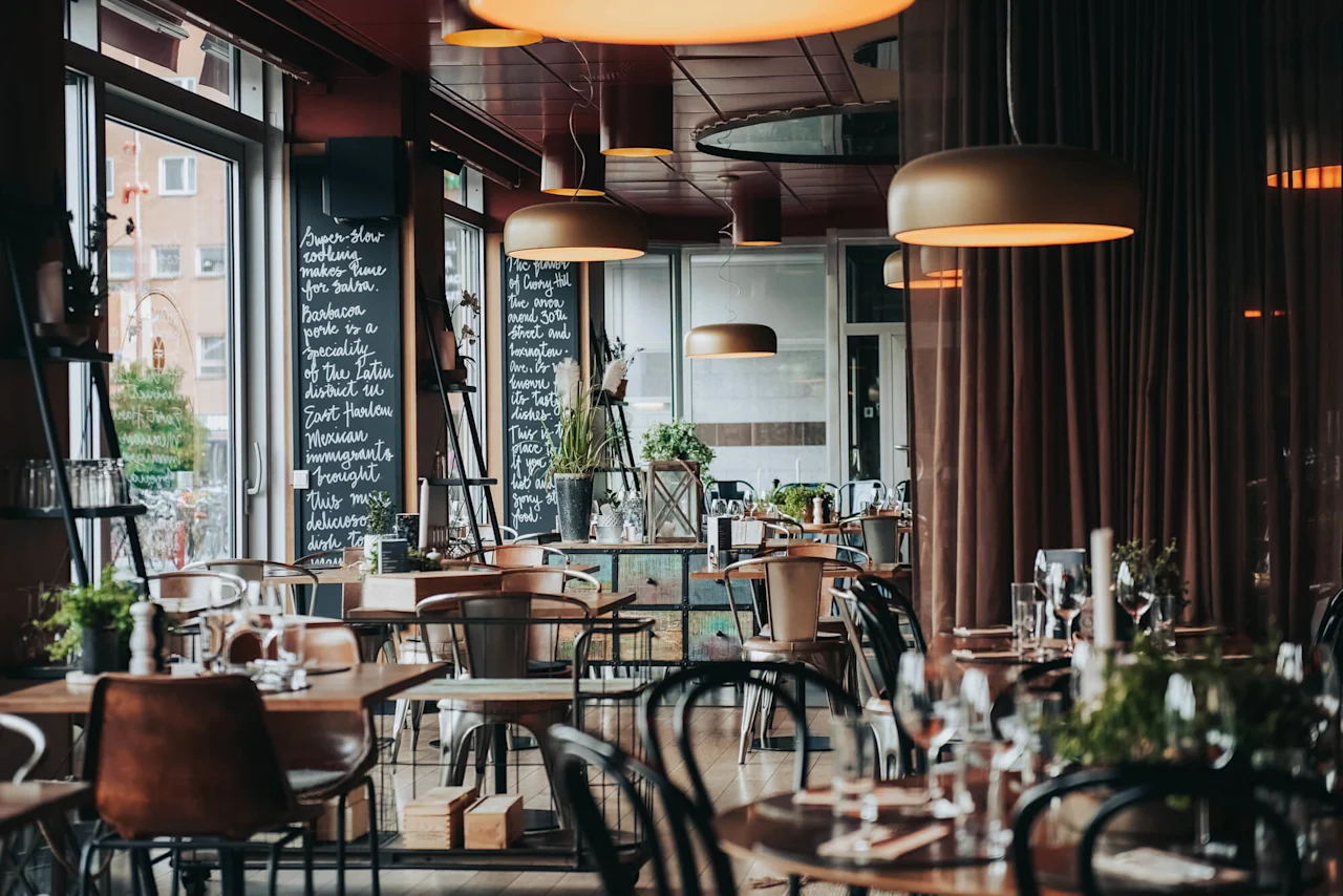 Elegant restaurant med treborde dekket for middag, omgitt av store vinduer og tavler med håndskrevet tekst. Myke lysarmaturer henger fra taket og skaper en koselig atmosfære.