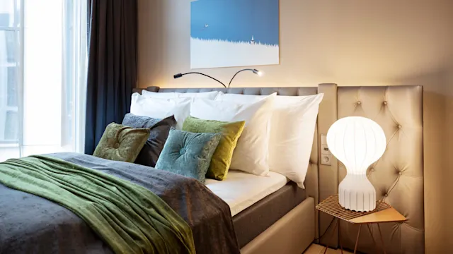 Bed adorned with white sheets and colorful cushions, positioned under a blue abstract painting. A modern lamp stands on a bedside table. Bright window with sheer curtains nearby.