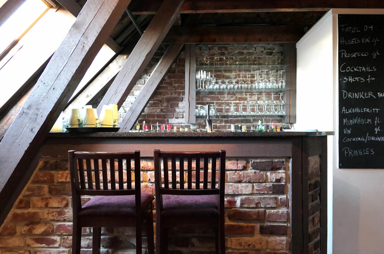 Two chairs face a bar counter under rustic wooden beams, with shelves of glasses and drinks; a menu board details available beverages.