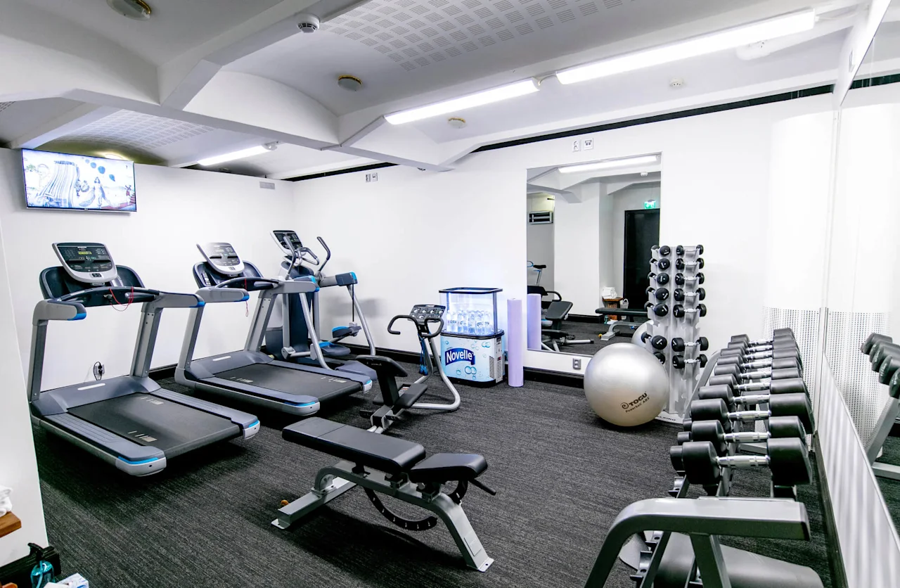Treadmills and exercise machines stand inside a gym with white walls, mirrors, and a row of dumbbells. A TV on the wall broadcasts visuals. "Novelle" water dispenser and an exercise ball are present.