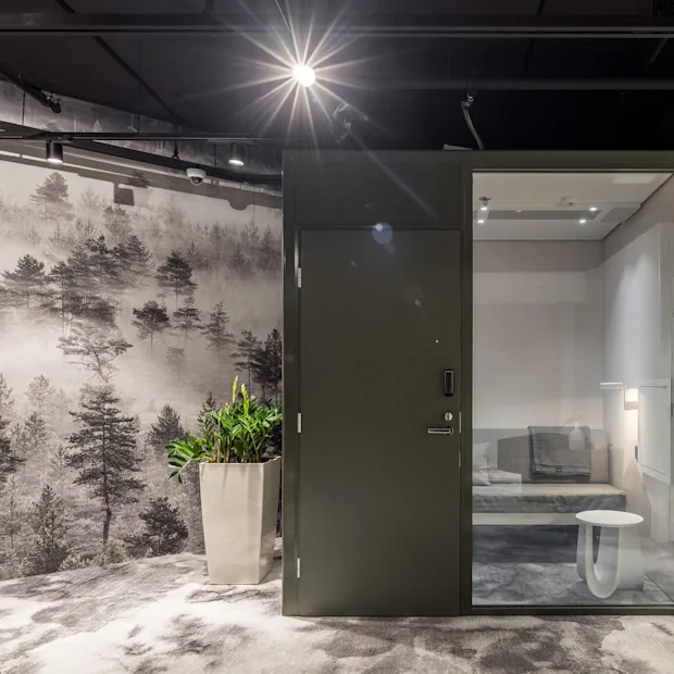 En minimalistisk kontorplass med et glassvegget møterom med en grå sofa og små bord, omgitt av et skogstema veggbilde og en stor plante i en potte.