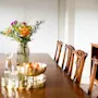 A vase with colorful flowers stands on a wood dining table, flanked by chairs in a bright room.