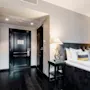 A neatly made bed with white pillows and dark headboard sits beside two stylish lamps in an elegant hotel room with dark wooden floors and light-colored walls.