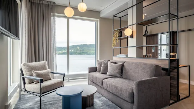 En grå sofa står i en moderne stue med stort vindu med utsikt over vann og åser, komplettert av en stilig lenestol, rundt bord og hengende lys.