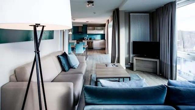 Et moderne oppholdsrom har en gulvlampe som lyser opp en beige sofa med blå puter. I nærheten står et salongbord og en flatskjerm-TV i et rom med store vinduer.