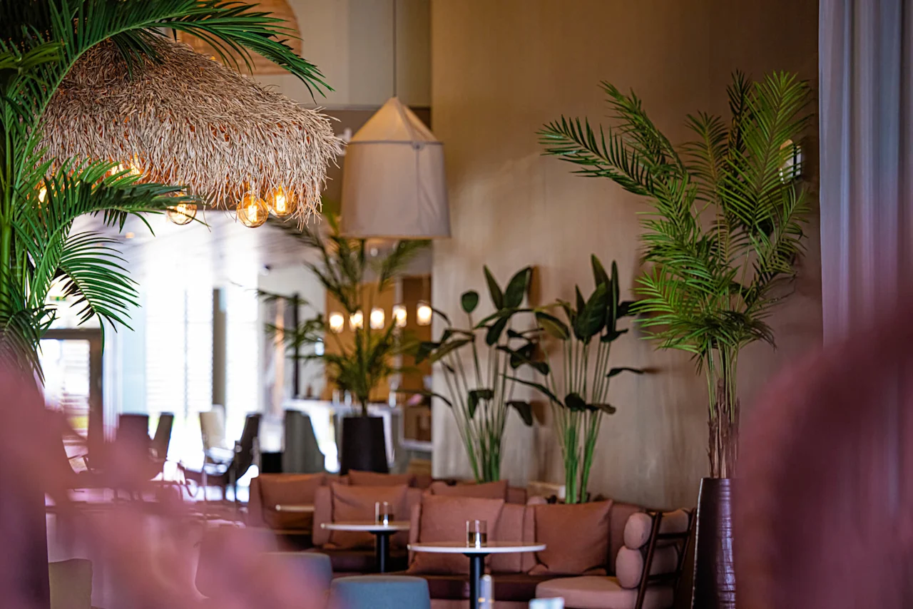 Myke sitteplasser er arrangert i en varmt opplyst lounge pyntet med tropiske planter og en taklampe i stråtak. Runde bord med drikkevarer skaper en koselig, innbydende atmosfære.