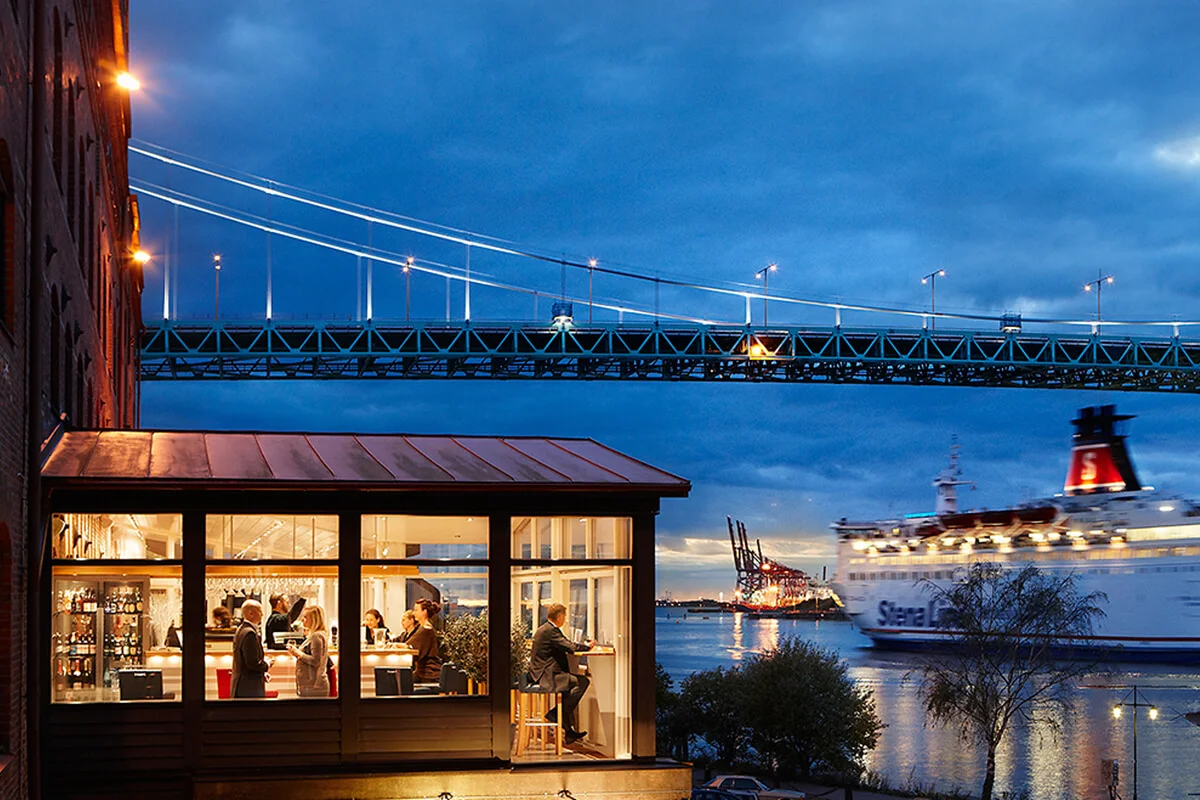 Ravintolavieraat ruokailevat lasiseinäisessä rakennuksessa joen rannalla. Silta kulkee yläpuolella, kun suuri "Stena Line" -lautta lipuu ohi iltavalossa.