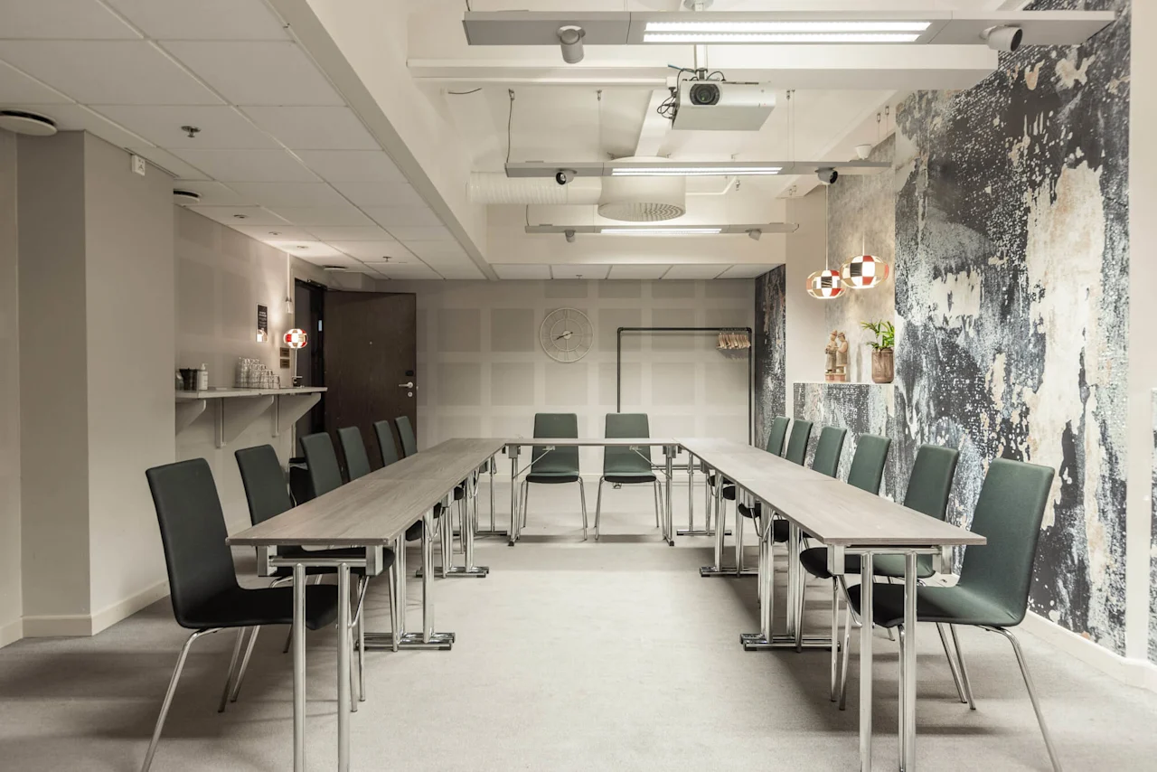 Møterom med U-formet bordoppsett, grønne stoler og moderne innredning inkludert mønstrede vegger og hengende lamper. En hylle har glass, og en takmontert projektor er synlig.