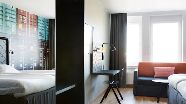 Ett hotellrum har två sängar, ett skrivbord med lampa och en soffa mot en vägg med stora fönster. Inredningen inkluderar en väggmålning av färgglada fraktcontainrar.