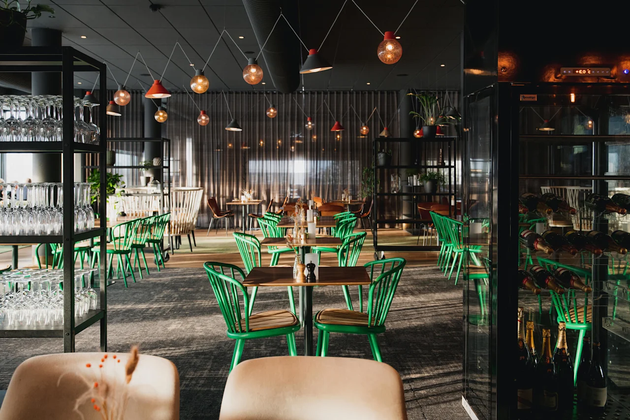 Bord med grønne stoler er plassert, omgitt av hyller med glass. Hengelamper henger over i en svakt opplyst moderne restaurant. En vinutstilling er på høyre side.