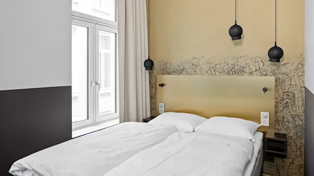 En omsorgsfullt bäddad säng med vita sängkläder står under en gul vägg med graffiti, i ett modernt rum prytt med två hängande svarta lampor och stora fönster med beige gardiner.