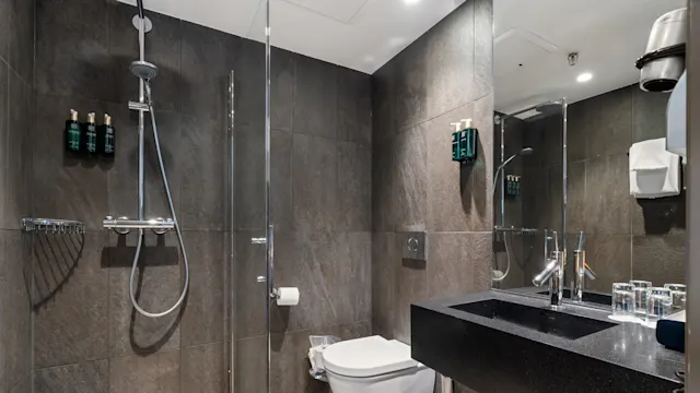 A modern bathroom features a glass-enclosed shower with toiletries, a wall-mounted toilet, and a black countertop sink. Dark tiles cover the walls, complemented by a row of glasses and a hairdryer.