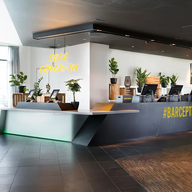 Resepsjonsdisk med dataskjermer; planter og neonskilt over som lyder "SELF CHECK-IN". Langt barområde med stoler, merket "#BARCEPTION", lyser grønt. Moderne interiør.