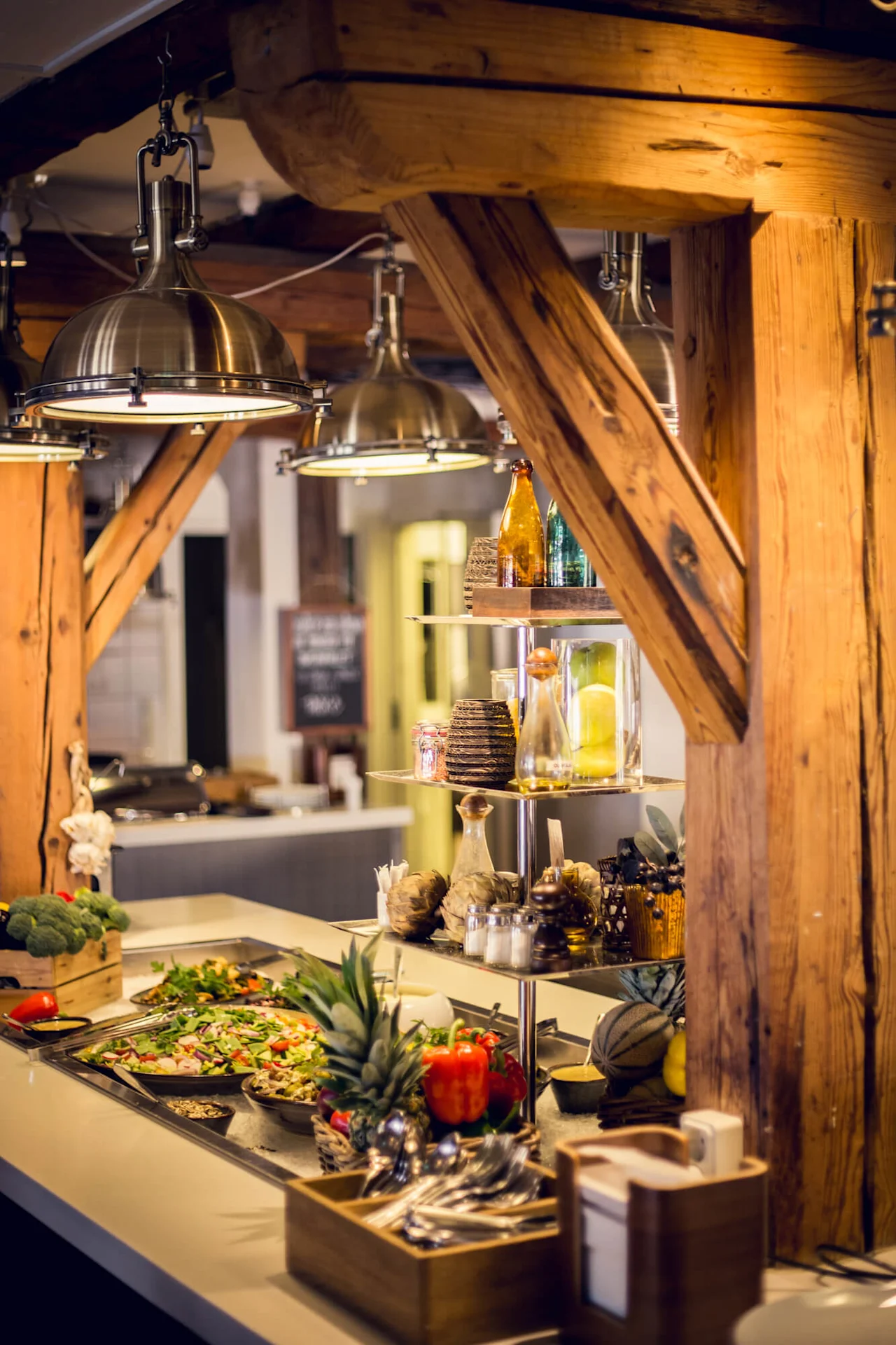 A buffet spread with salads, fruits, and cakes under metal lamps, framed by wooden beams inside a cozy dining area.