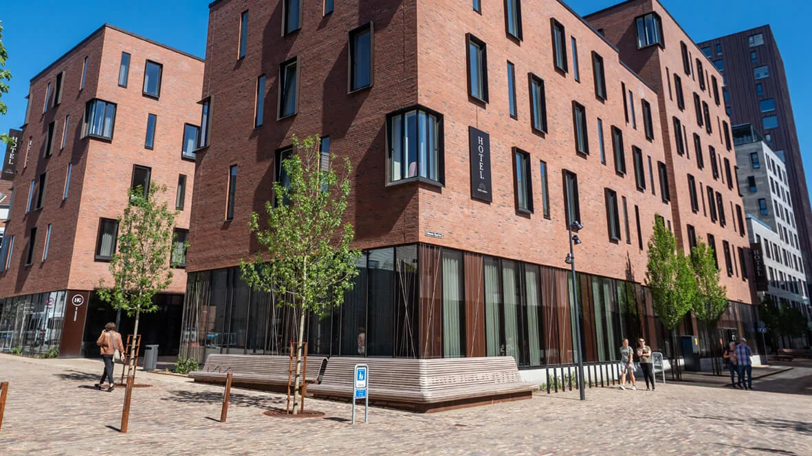 En modern, flervånings tegelbyggnad som är ett hotell med stora fönster ligger vid en gata med träd och bänkar. Människor promenerar i närheten. Text: "HOTEL".