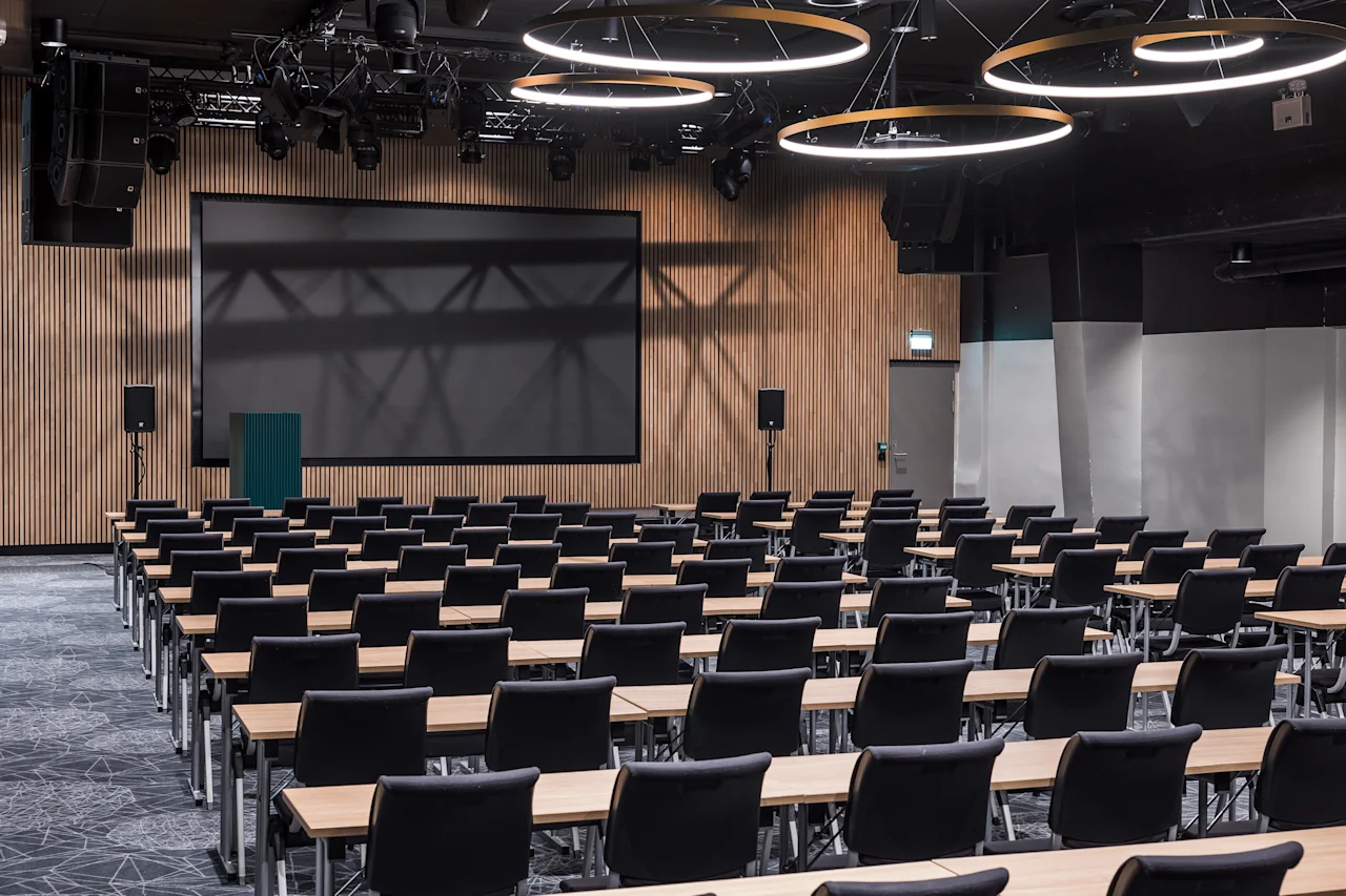 Konferanserom med rader av bord og stoler vendt mot en stor skjerm på en vegg med trepanel. Sirkulære lamper henger fra taket og lyser opp rommet.