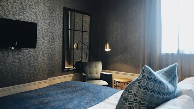 Seng dekket med blått teppe, i et moderne rom, nær en mønstret lenestol og gulvlampe i gull. Stort speil og flatskjerm-TV på tapetserte vegger; vindu med tynne gardiner.