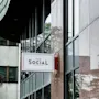 A rectangular sign shows "THE SOCIAL BAR & BISTRO" in gold letters, attached to a building with reflective glass and textured stone panels, surrounded by other urban structures.