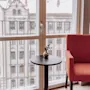 A red armchair and small table by a large window overlooking European architecture.