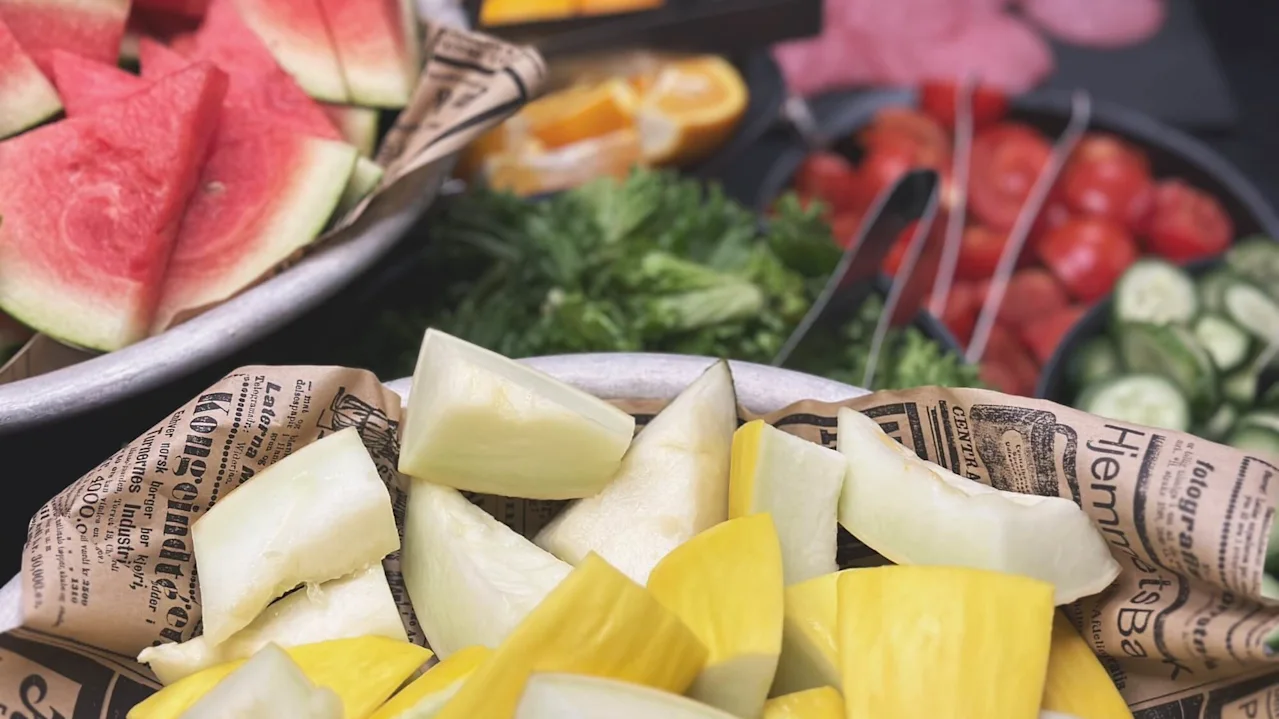 Vannmelon og blandede grønnsaker ligger i boller på et bord, omgitt av skiver av agurk, tomater og grønnsaker. Bakgrunnen har flere skivede frukter og kjøtt. Avispapir er synlig.