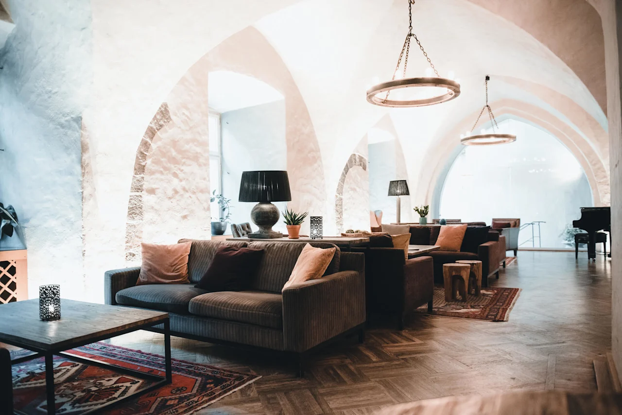 Cozy seating area with sofas and cushions, featuring wooden tables and decorative lamps, under arched ceilings. Plants and a grand piano enhance the elegant, serene atmosphere.