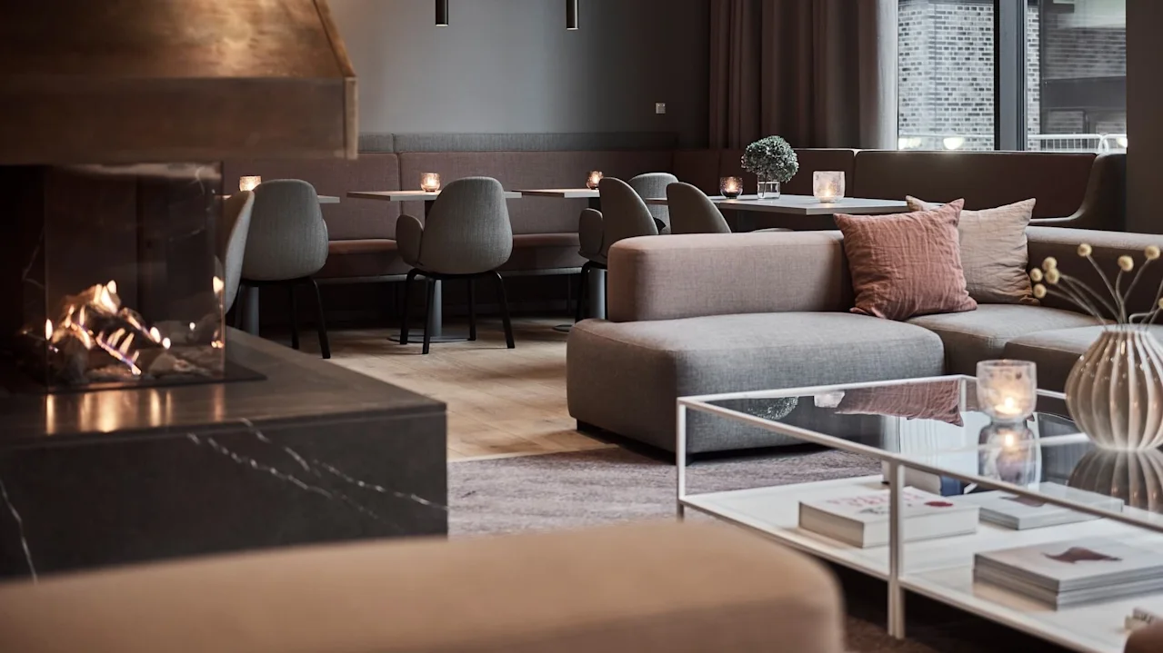 Modern lounge med eldstad, grå soffor och ett stilrent soffbord. Ljus och minimalistisk inredning ger en mysig atmosfär med stora fönster i bakgrunden.