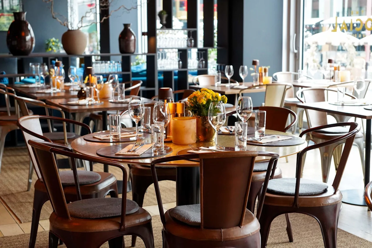 Et dekket bord med blomster og glass, arrangert på trestoler i en koselig, varmt opplyst restaurant med store vinduer og dekorative vaser i bakgrunnen.