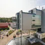 En moderne bygning med glass- og metallelementer strekker seg over et basseng. Uteområder med sitteplasser og planter omgis av trær og bygninger med røde tak.