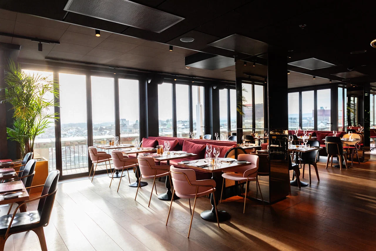 A bright, modern restaurant with wooden floors features pink and black chairs around set tables. Large windows provide a city view, and lush green plants enhance the elegant atmosphere.