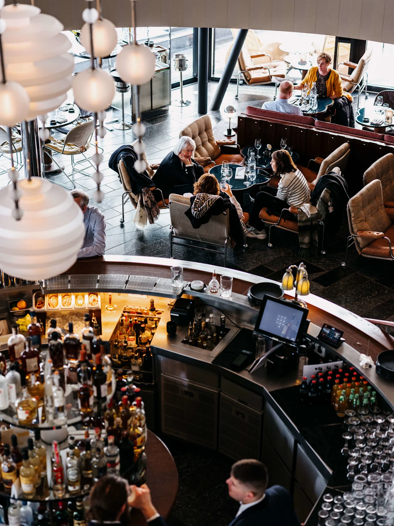Gjester spiser og sosialiserer i en elegant restaurant med flere nivåer, hengende globelys, plysjseter og et fremtredende barområde.