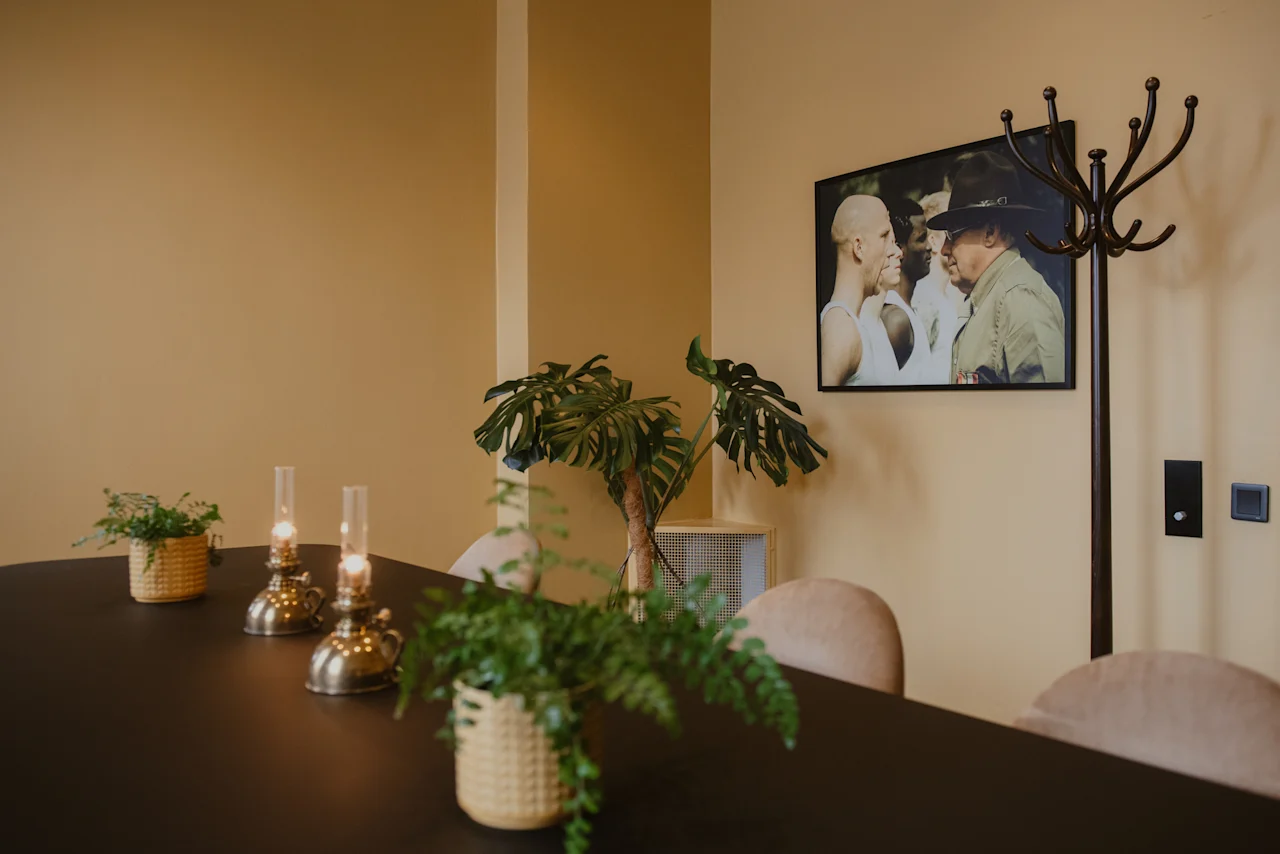 Et svart bord har to tente oljelamper og potteplanter. I nærheten henger et innrammet bilde på en beige vegg. En stumtjener står ved siden av bordet.