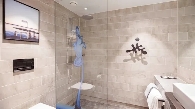 En dusj slipper ut blått vann i et moderne bad. Flislagte vegger og en glassdør rammer inn området. Håndklær henger ved siden av en hvit vask. Kunst pryder en vegg.