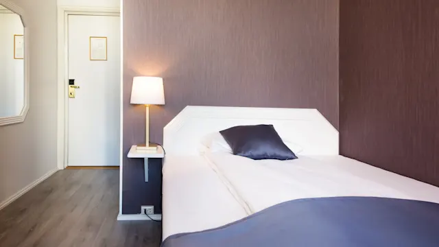 En enkeltseng med hvite og blå sengetøy, ved siden av en tent lampe, i et rom med brune vegger og et speil som reflekterer inngangsdøren.