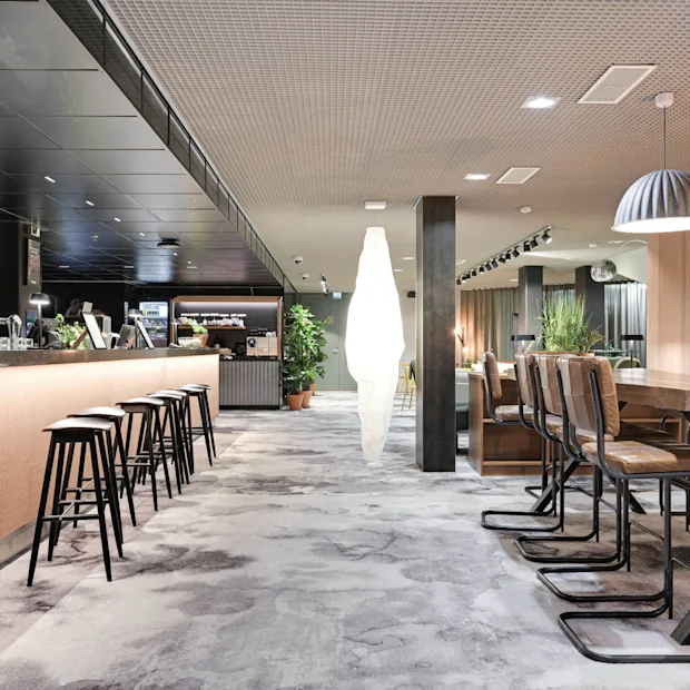En moderne hotellsalong har en elegant bar med høye krakker, et langt felles bord og stilig dekor. Planter gir grønt miljø. Tekst på veggen: "COMFORT HOTEL XPRESS."