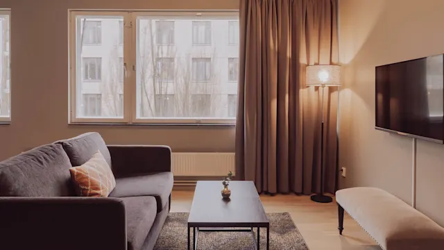 A gray sofa with an orange cushion faces a black coffee table in a living room. Nearby, a lamp stands beside floor-length curtains, opposite a wall-mounted TV.