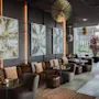 Hyggelig lounge med brune sofaer og mønstrede puder, abstrakt kunst på mørke vægge, og runde borde. Unikke pendellamper hænger over, og store vinduer viser en have.