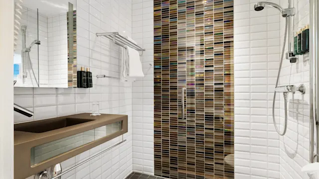 Bathroom with a brown sink, glass partition, and white tiled walls. A towel is on a metal rack, and a shower with a hose and bottles is nearby.