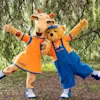 A giraffe and a bear costume character dance playfully, wearing orange and blue outfits in a park. Text on outfits: "Lollo" and "Bernie." Background features leafy trees and grass.