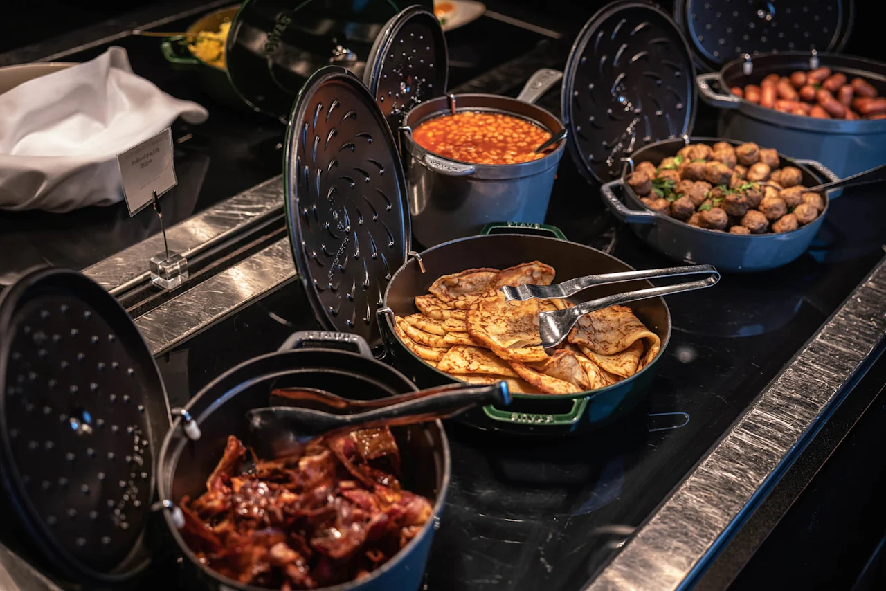 Pannekaker ligger i en panne med tang på en buffet. I nærheten er bakt bønner, pølser og bacon. En tildekket skål har et lite skilt med teksten "Lepeske Blinis/Crepes."