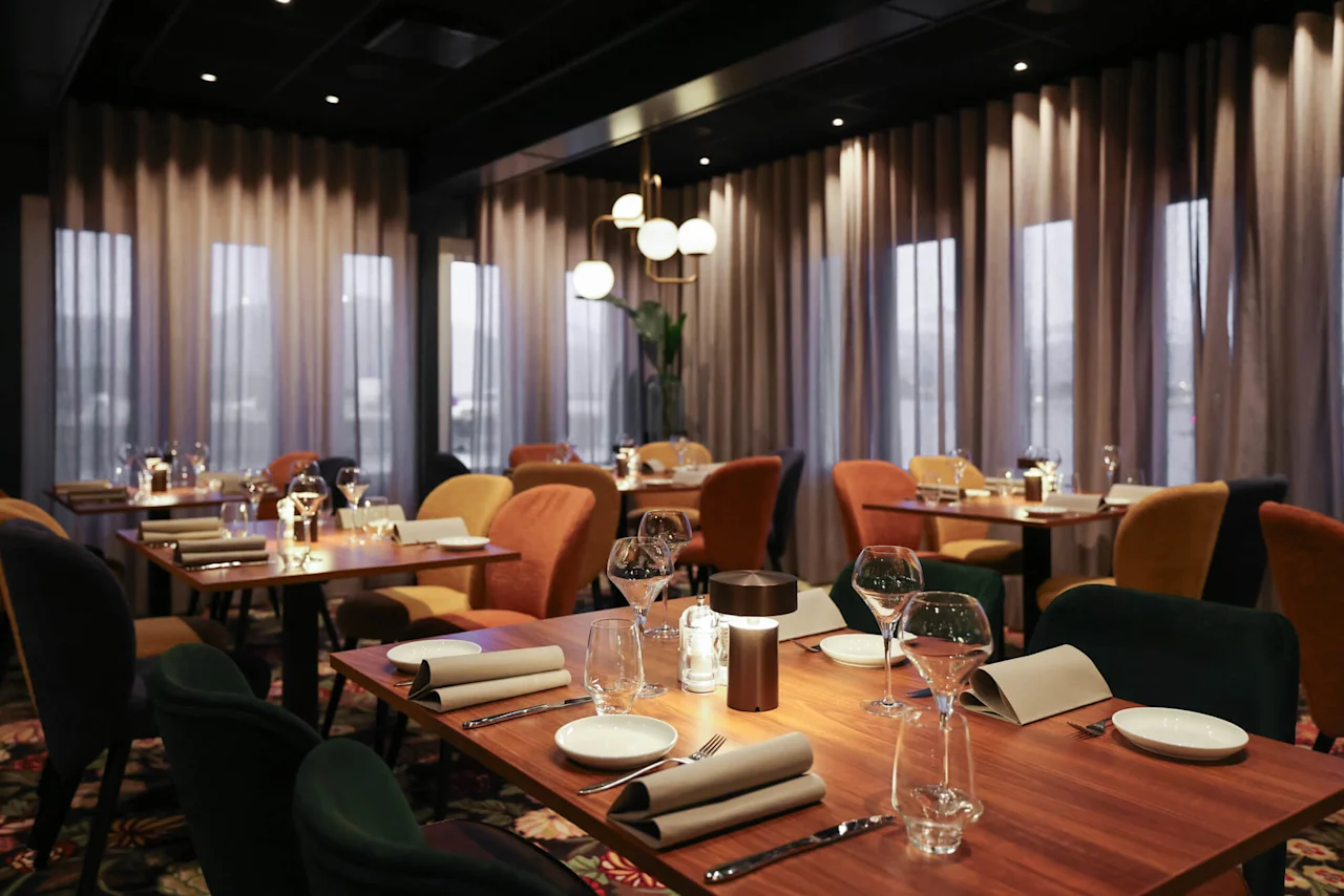 En restaurantinteriør med pænt dækkede træborde, vinglas og foldede servietter. Blødt lys og afdæmpede gardiner skaber en intim atmosfære.