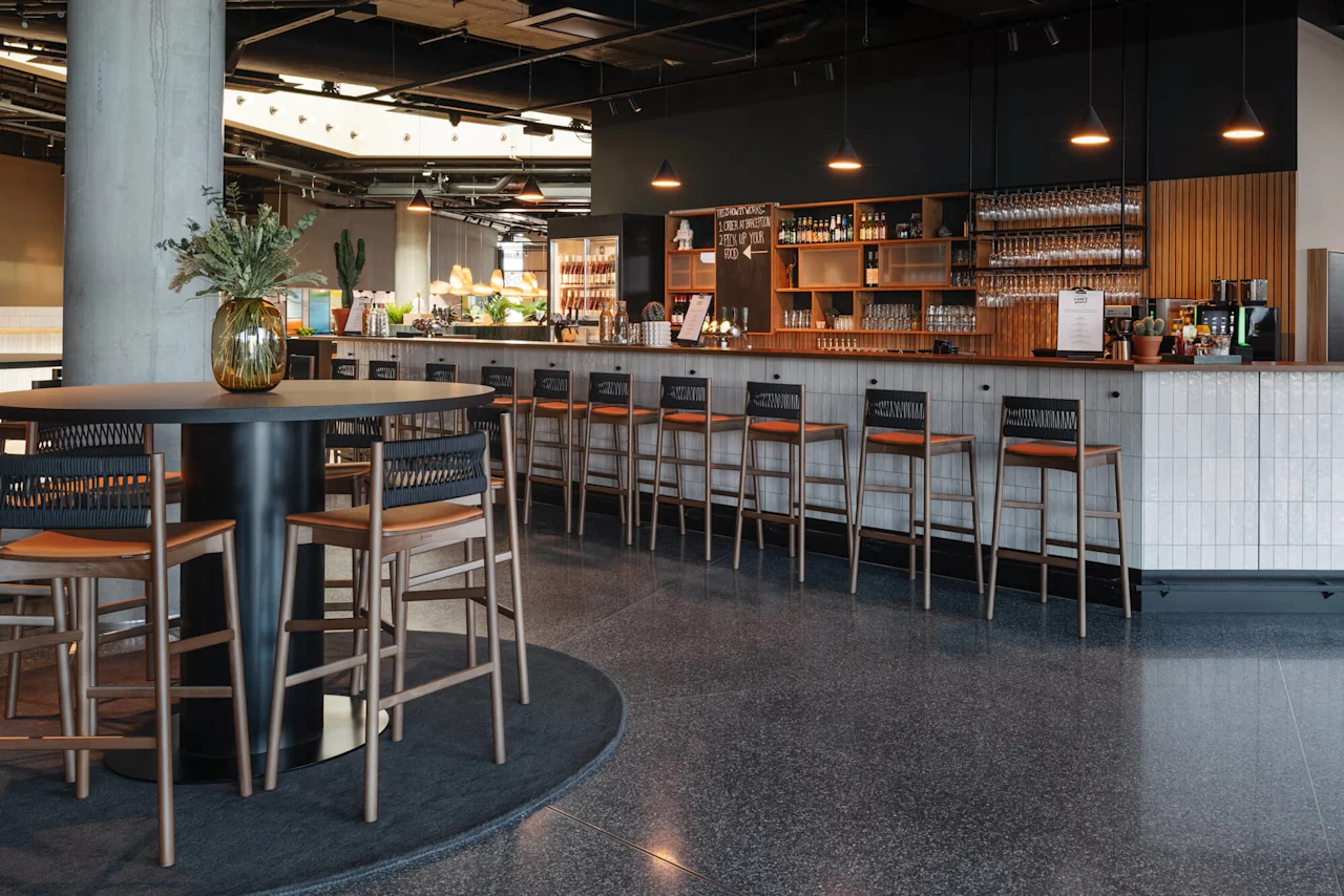 A modern bar with tall chairs lined along a counter, lit by pendant lights. A round table with plants is in the foreground.