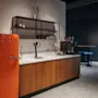 A retro orange SMEG refrigerator stands beside a modern kitchen counter with a marble top, shelves with dishes, a coffee dispenser, and a tray of snacks in a stylish interior.